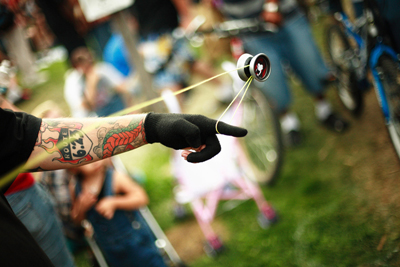 A figura mostra o braço esquerdo de um homem com marcas de tatuagem e usando uma luva. Ele está circulando um brinquedo de ioiô, que está no ar e conectado pela corda à sua mão. Algumas pessoas estão em segundo plano assistindo ao truque do ioiô.