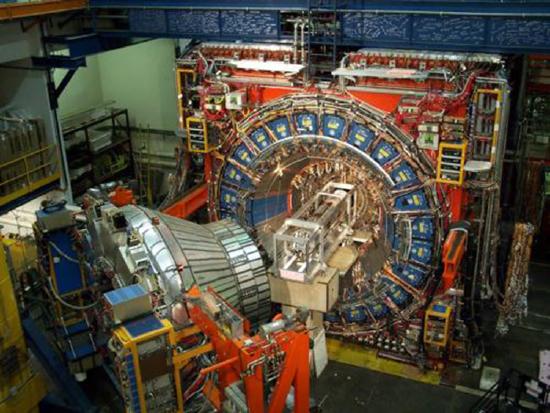 The image shows the picture of a huge cylindrical shaped proton decay detector with its main door open. It is as high as a double decker bus and as long as a small house. An untold number of cables, wires, and detector modules are arranged in a cylinder around a rectangular crate-like object containing another smaller cylindrical object.