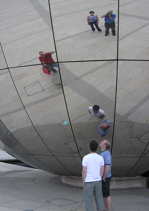 people are shown standing beneath a giant mirrored sphere While looking at their reflections.