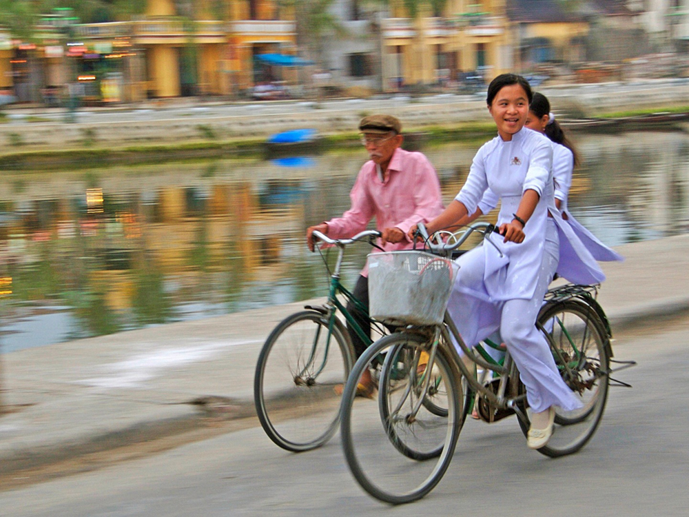 Três pessoas andando de bicicleta ao longo de um canal. Os edifícios borrados ao fundo transmitem uma sensação de movimento dos ciclistas.