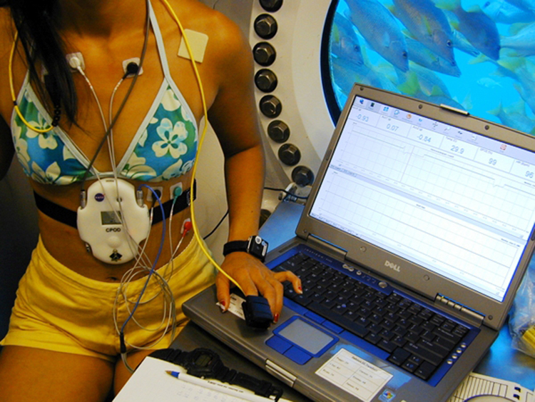 Fotografia de uma cientista da NASA em um habitat subaquático registrando seus sinais vitais usando um dispositivo portátil e um laptop.