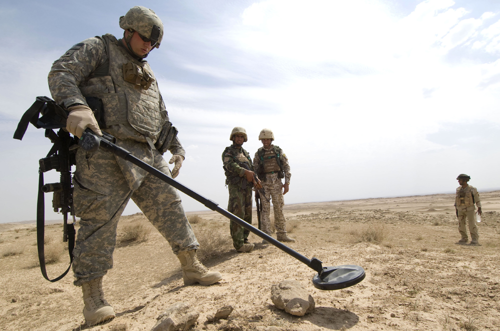 ] Fotografia de vários soldados em campo aberto. Um soldado está procurando por explosivos escaneando a superfície usando um detector de metais.