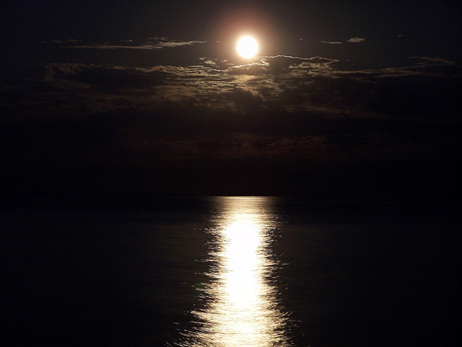 Uma noite escura é iluminada pela luz da lua. A luz da lua está caindo sobre o lago e, à medida que atinge, a superfície brilhante do lago a reflete. Uma faixa brilhante de luar é vista refletindo do lago em um fundo escuro refletindo o céu noturno.