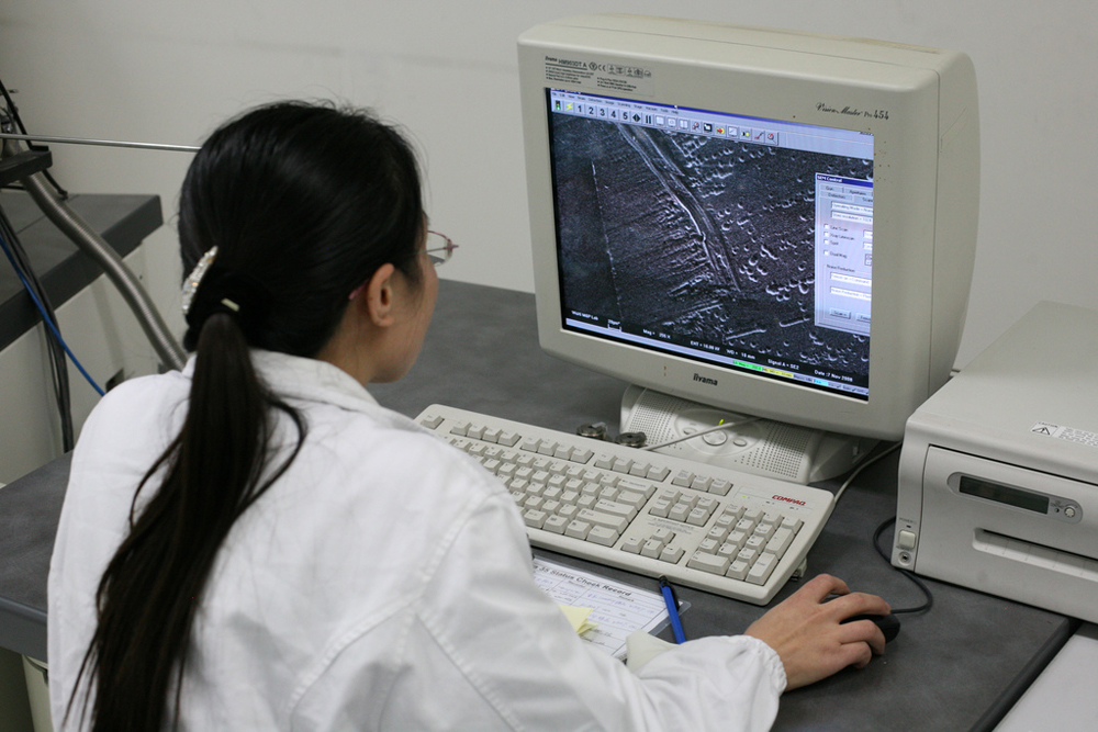 A imagem mostra a vista frontal de um computador desktop junto com um teclado e uma impressora com uma imagem sendo exibida na tela do computador. Também mostra as costas de uma mulher segurando o mouse.