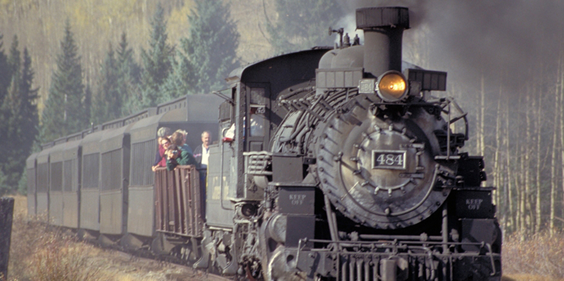 Uma máquina a vapor e vários vagões de passageiros são mostrados viajando pelos trilhos de um trem. O trem tem algumas pessoas a bordo.