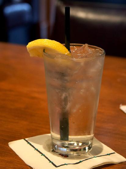 Photograph shows a glass of a beverage with ice cubes and a straw, placed on a paper napkin on the table. There is a piece of sliced lemon at the edge of the glass. There is condensate around the outside surface of the glass, giving the appearance that the ice is melting.