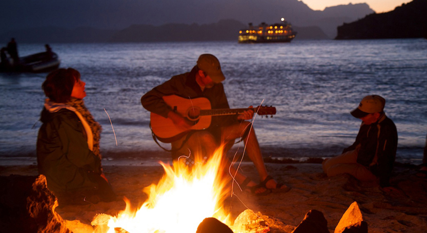 Na figura, um casal e seu filho estão sentados ao lado de uma praia à noite, ao redor de uma lareira acesa a lenha. O homem está tocando violão.