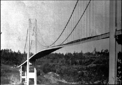 A figura mostra uma foto em preto e branco da Ponte Tacoma Narrows, da vista lateral esquerda. O meio da ponte é mostrado aqui em um estado oscilante devido aos fortes ventos cruzados.