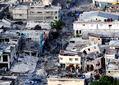 A destruição causada por um terremoto em Porto Príncipe, Haiti. Alguns edifícios são mostrados nos dois lados de uma rua. Dois edifícios estão completamente destruídos. Pessoas de resgate são vistas ao redor.