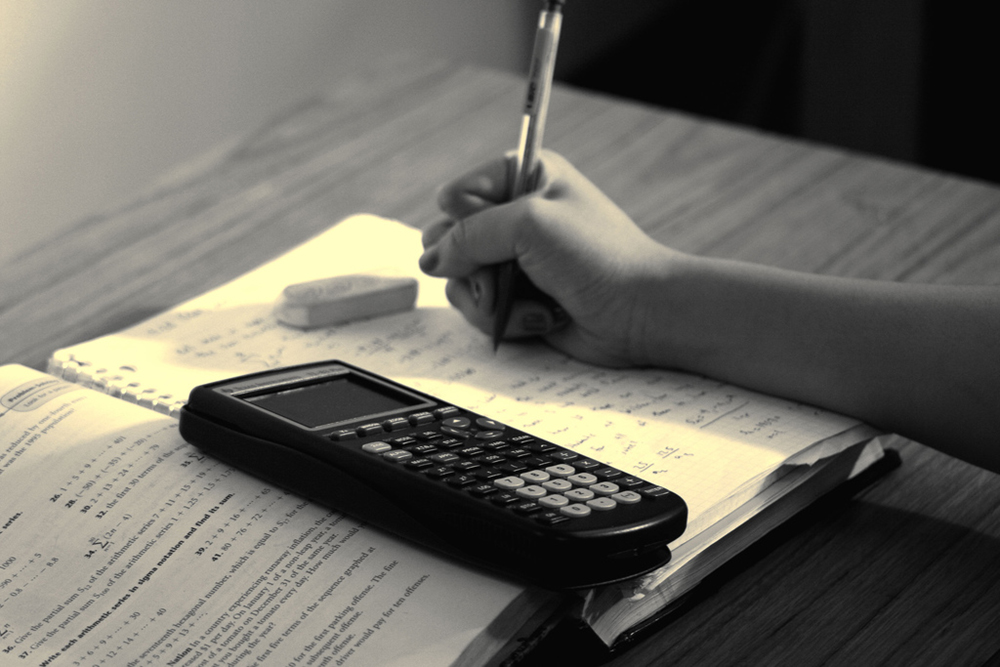 Foto em close-up de uma mão escrevendo em um caderno. No topo do caderno há uma calculadora gráfica.