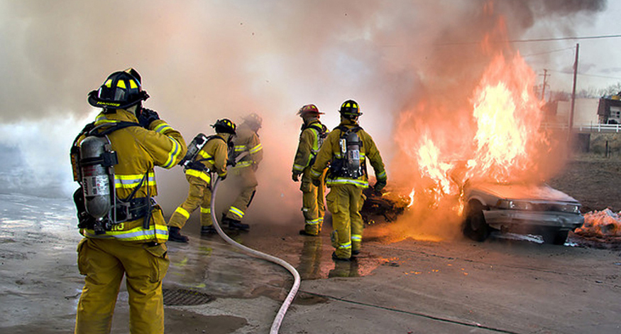 A fotografia mostra um grupo de bombeiros uniformizados usando uma mangueira para apagar um incêndio que está consumindo dois carros.