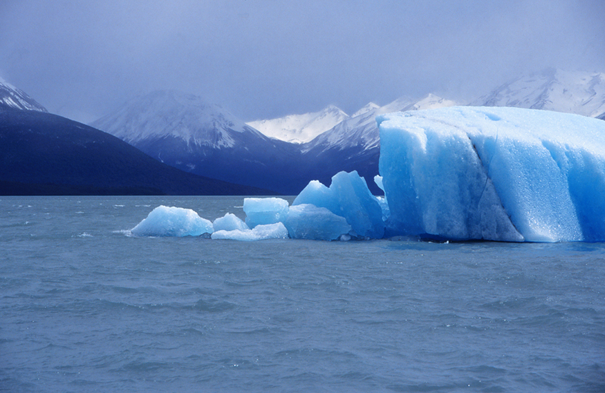 A figura mostra alguns icebergs de cor azul flutuando na água sob montanhas cobertas de neve e um céu nublado. Alguns dos icebergs na frente estão derretendo.