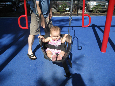 In the figure shown, a small child is seated in a spring swing, tied with a belt at his waist. In the back is his father, who is pushing the swing in the to and fro motion.