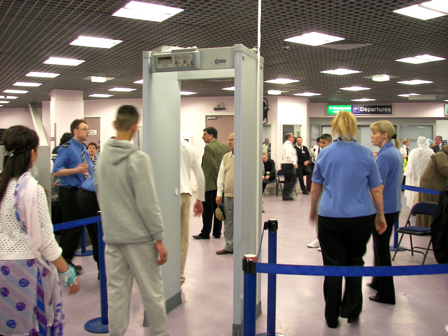 Fotografia de pessoas ao redor de um portão de segurança no terminal de embarque do aeroporto.