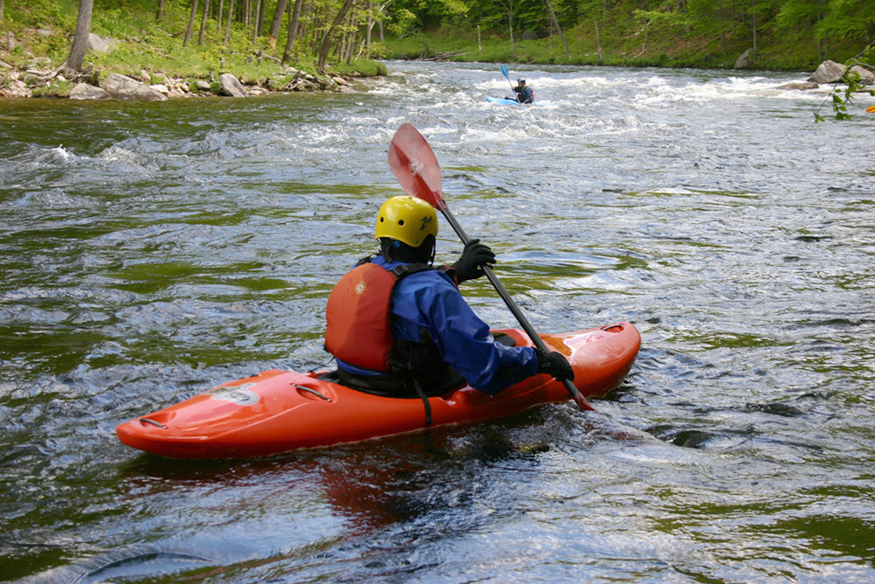 Un hombre con remo en la mano está haciendo kayak río abajo en un río poco profundo que fluye rápidamente.