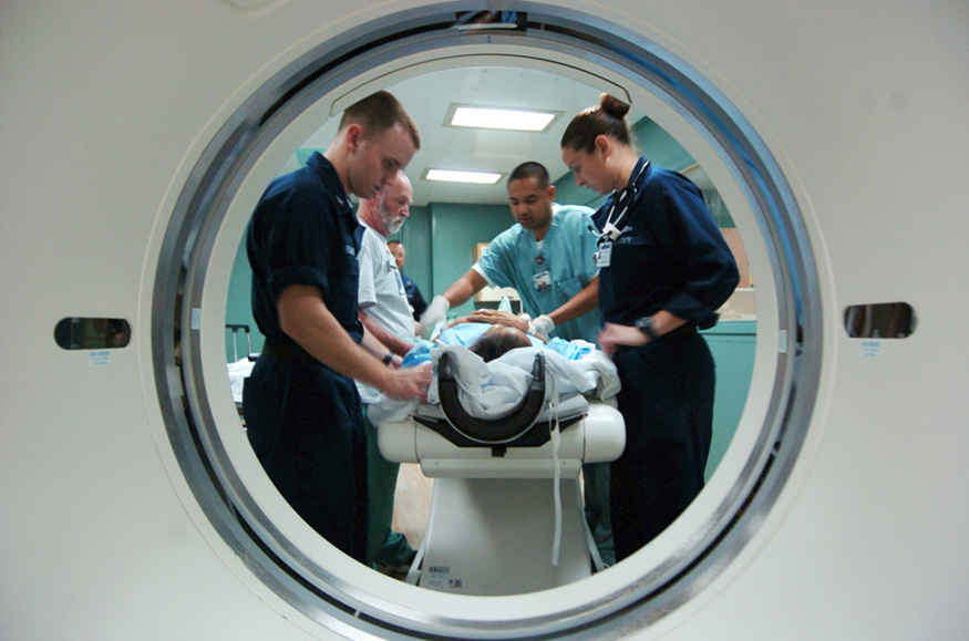 Uma imagem fotográfica tirada pela porta de um scanner C T, mostrando um paciente em uma maca cercado por três equipes de enfermagem e um médico que estão fazendo a tomografia computadorizada do paciente.