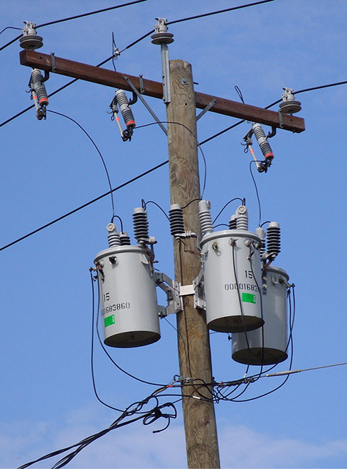 Photographie de transformateurs sur un poteau électrique. Il y a trois transformateurs, chacun placé dans un récipient cylindrique.
