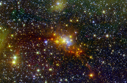 Uma imagem telescópica mostrando várias estrelas. Um enxame brilhante no centro tem estrelas amarelas, alaranjadas e azuis.